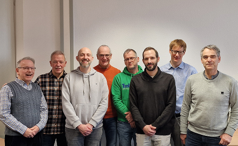 Eight men standing indoors against a plain wall, dressed casually in sweaters, hoodies, and shirts.