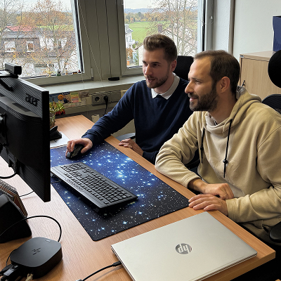 Two men working together at a desk, looking at a computer screen with a keyboard and mouse on a space-themed mousepad, a closed HP laptop nearby.